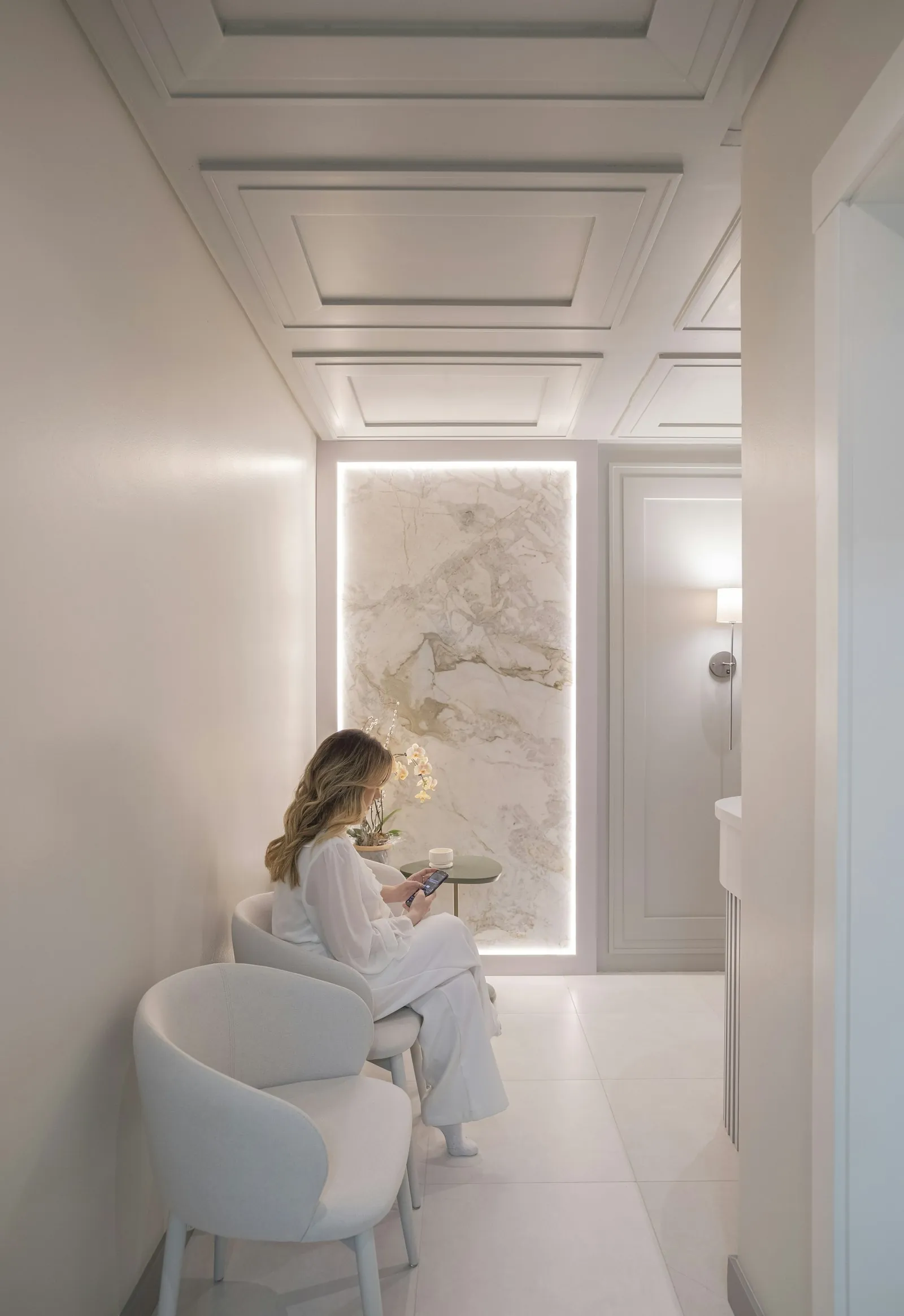 The waiting area of a private consulting practice: a curved armchair beside a backlit marble panel in a bright, warm-white room. A representative interior photograph; it does not show either clinic.